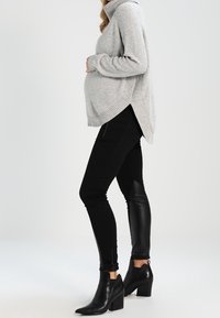 Grau gestreifter Strickpullover mit hohem Kragen und asymmetrischem Saum, kombiniert mit schwarzen Hosen mit Kunstleder-Akzenten und hochhackigen Stiefeletten.