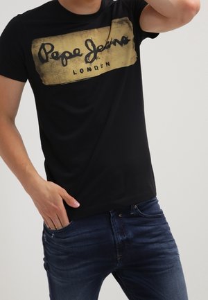 Mann trägt ein schwarzes Pepe Jeans London T-Shirt mit goldenem Logo und dunkelblaue Jeans, eine Hand in der Tasche und die andere erhoben.