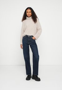 Camisola de malha bege claro com gola alta, jeans azuis de perna reta com design clássico de cinco bolsos e botas pretas com plataforma.