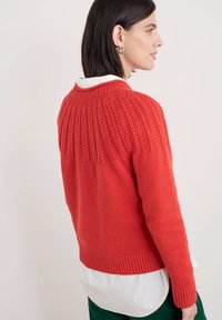 Röd stickad tröja med en zigzag-texturerad yoke, ribbade ärmslut och fåll, bärs över en vit skjorta med krage. Mjuk och figursydd design.