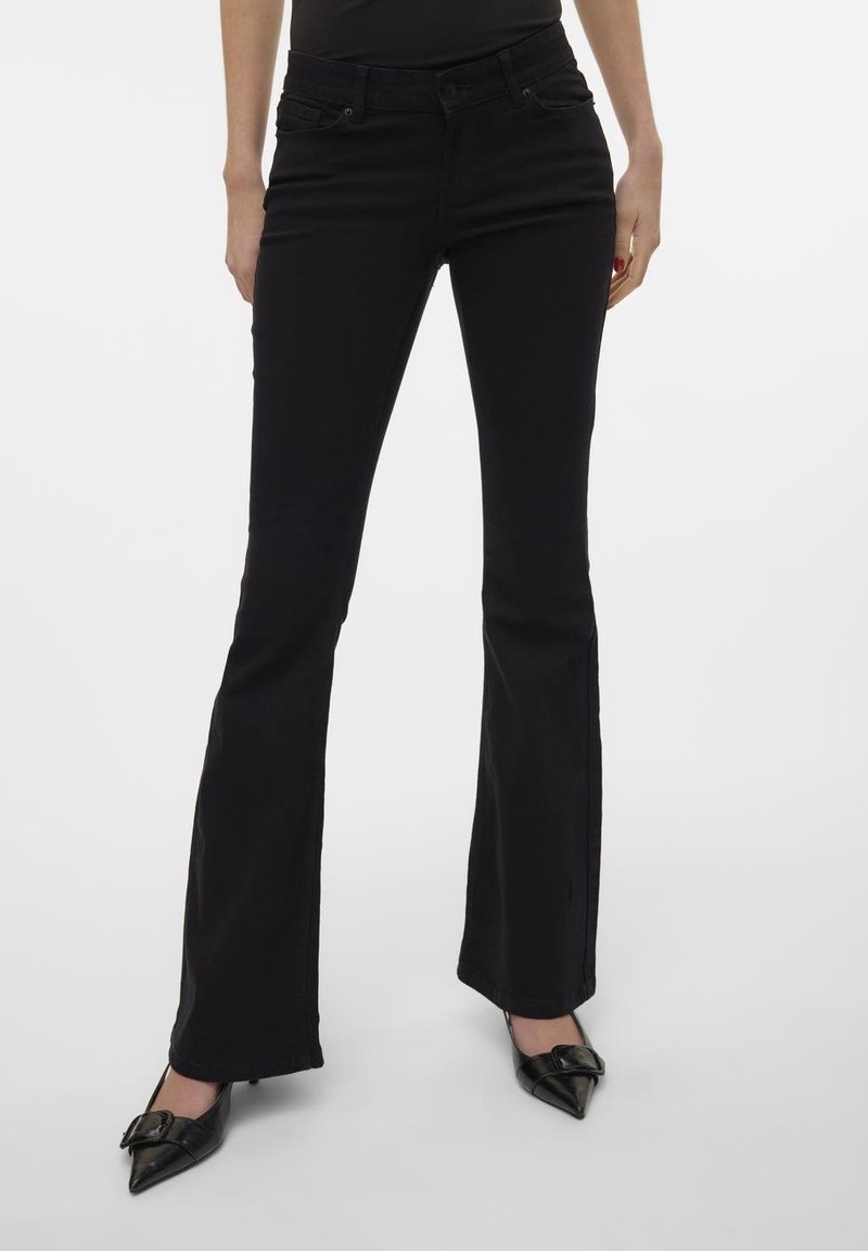 Jean noir taille haute coupe bootcut porté par une personne debout, associé à des talons noirs pointus avec des détails en nœud.