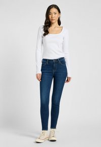 Haut à manches longues blanc avec un décolleté carré associé à un jean skinny bleu marine. L'ensemble comprend des baskets montantes beiges avec des accents rouges.