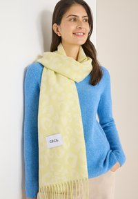 Écharpe jaune pâle avec motif léopard, bords frangés, et une étiquette portant la mention "CECIL", portée sur un pull bleu côtelé.