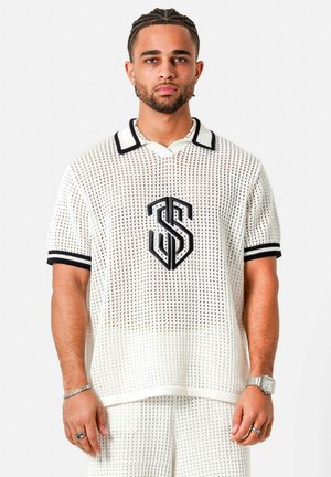 Man met gevlochten haar draagt wit mesh shirt met korte mouwen en broek met zwart logo, staand tegen een effen lichte achtergrond.