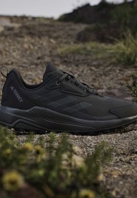 Zwarte Adidas Terrex trail hardloopschoen op een rotsachtig buitenpad met groen op de voorgrond en achtergrond.