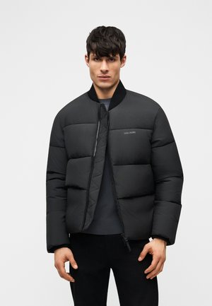 Homme portant une veste matelassée noire avec une fermeture éclair, debout devant un fond gris clair uni, faisant face à l'avant.