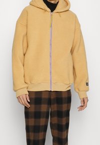 Osoba ubrana w beżowy hoodie z zamkiem błyskawicznym typu sherpa, z żółtymi sznurkami i brązowymi oraz czarnymi spodniami w kratę, stoi na jednolitym tle.