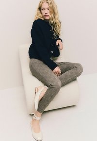 Žena s dlhými kučeravými blond vlasmi, oblečená v čiernom svetríku na gombíky, kostkovaných nohaviciach a bielych topánkach na členkovom remienku, je posadená na texturovanom krémovom kresle.