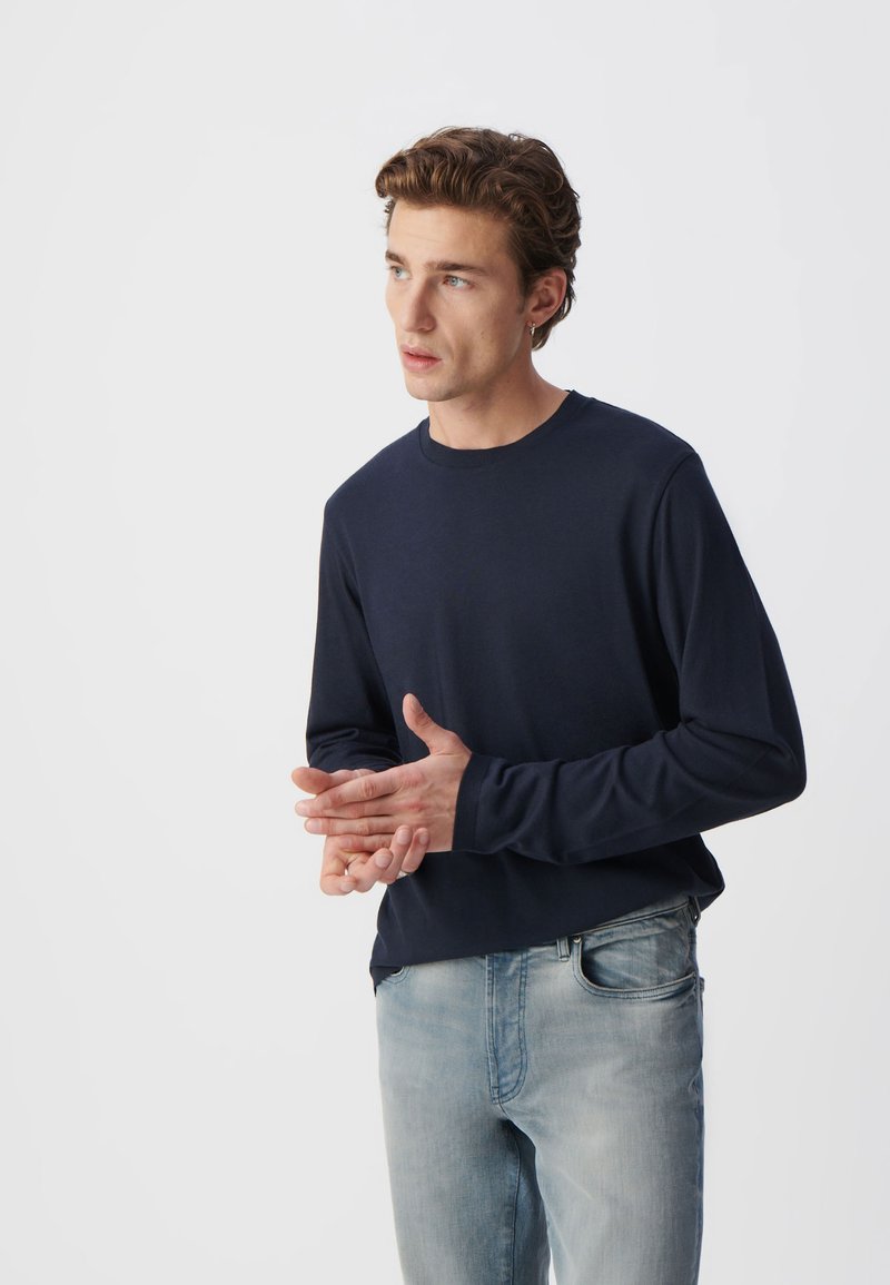 Marineblaues Langarmshirt aus weichem Stoff, runder Halsausschnitt, kombiniert mit hellblauen Jeans. Das Model steht mit gestikulierenden Händen.