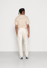 Camiseta de algodón beige con texto gráfico en la parte posterior, combinada con pantalones blancos. Zapatillas en blanco y negro completan el conjunto.
