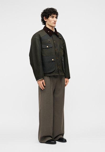 Νέος άνδρας με σγουρά μαλλιά, φορώντας σκούρο πράσινο σακάκι με κουμπιά και πολλές τσέπες και φαρδιά γκρι παντελόνια, στέκεται πλάγια.