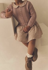 Pull en tricot rose avec une texture côtelée et un col, associé à une jupe beige à imprimé floral. Chaussures marron à lacets et chaussettes blanches à volants.