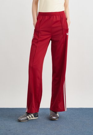 Persoon draagt rode Adidas joggingbroek met witte strepen, handen in de zakken, en zwarte Adidas sneakers, staand op een blauw tapijt.