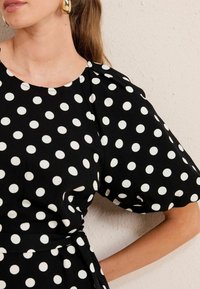 Rochie neagră cu buline mari albe, având mâneci scurte tip puff, decolteu rotund și un detaliu cu talie legată. Textură de țesătură netedă.
