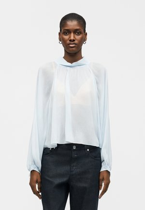 Γυναίκα που φορά ένα διάφανο γαλάζιο μπλουζάκι με μακρύ μανίκι, πιέτες και ψηλό γιακά, συνδυασμένο με σκούρο τζιν ψηλόμεσο.