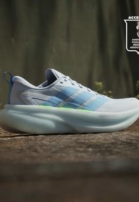 Бяла и синя маратонка Adidas с дебела омекотена подметка, поставена на открита земя на слънчева светлина.
