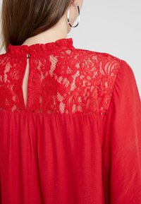 Rød bluse med en blonde ryg og nøglehul-design, rynket halsudskæring og blødt, flydende stof med en glat tekstur.