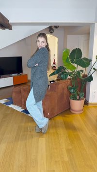 Pelēka adīta jaka, zili džinsi un pelēki zābaki uzvilkti iekštelpās. Brūna korduroja dīvāns un zaļa puķe dekorētā dzīvojamajā telpā.