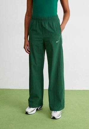 Personne portant un pantalon de survêtement large Nike vert foncé avec une taille élastique et des baskets blanches sur un sol vert et un fond blanc uni.