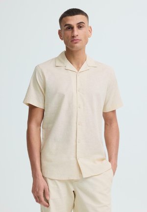 Man with short hair wearing a cream short-sleeve button-up shirt and matching light pants, standing with one hand in pocket.