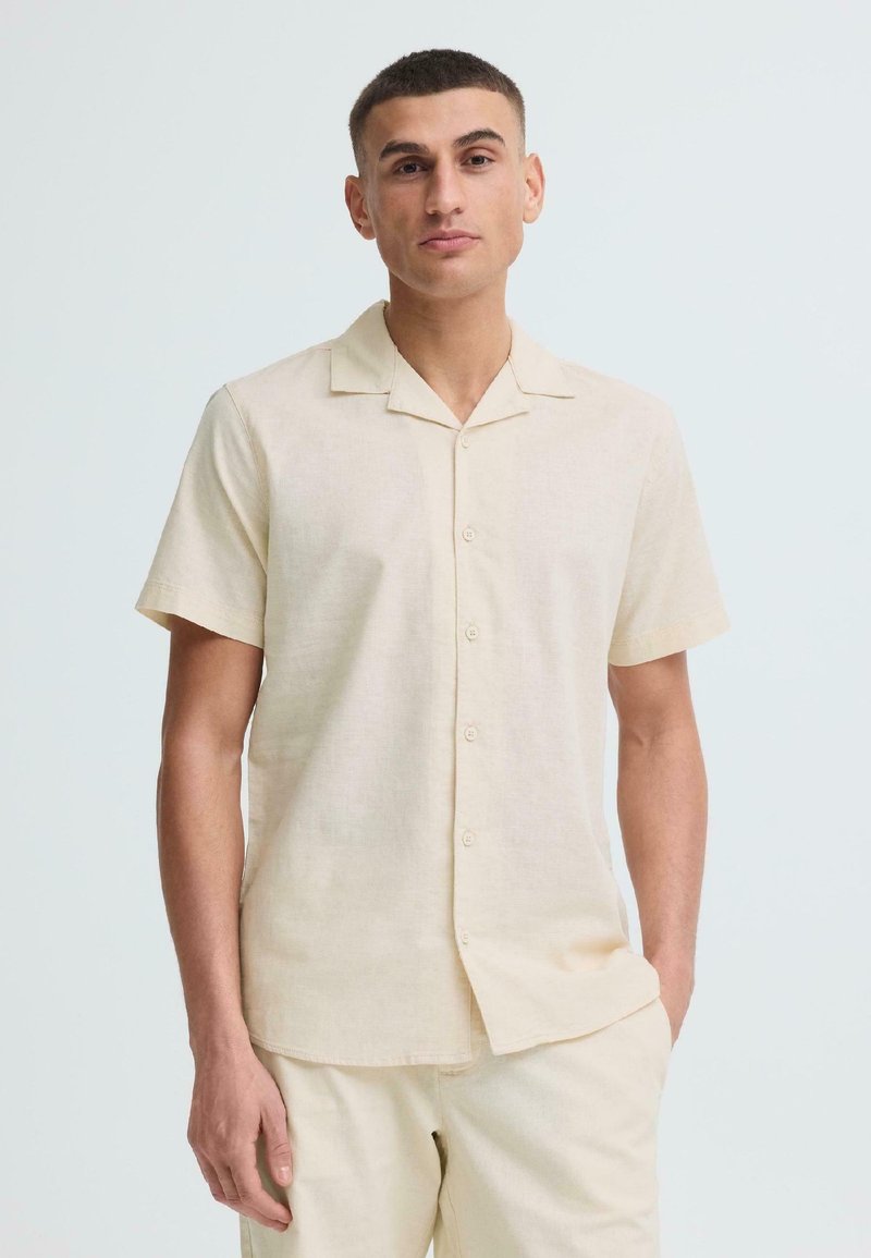 Homme aux cheveux courts portant une chemise crème à manches courtes boutonnée et un pantalon clair assorti, debout avec une main dans la poche.