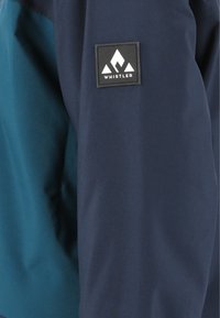 Miehen takki, jossa on laivastonsininen ja teal väri, valmistettu sileästä, vettä hylkivästä kankaasta, jossa on neliömäinen musta "Whistler" -logo.