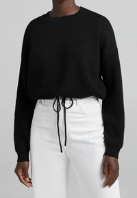 Schwarzer Strickpullover mit rundem Ausschnitt, Design mit fallenden Schultern und verstellbarem Kordelzug in der Taille, kombiniert mit weißen High-Waist-Jeans.