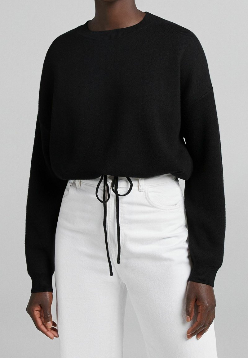 Schwarzer Strickpullover mit rundem Ausschnitt, Design mit fallenden Schultern und verstellbarem Kordelzug in der Taille, kombiniert mit weißen High-Waist-Jeans.