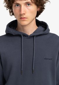 Sudadera con capucha de color azul marino, hecha de algodón suave. Cuenta con un bolsillo frontal y cordones con puntas de metal. Logotipo de "Element" bordado en un tono a juego.