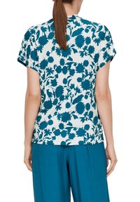 Korte mouw top met een teal bloemenpatroon op een witte achtergrond, ontspannen pasvorm, zachte stof, ronde halslijn en een subtiele naaddetail op de rug.