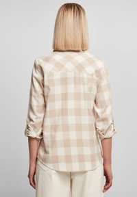 Personne aux cheveux blonds raides portant une chemise beige et blanche à carreaux à manches longues retroussées, vue de dos.