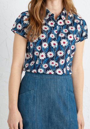 Kobieta nosząca granatową koszulę z krótkim rękawem z białym i czerwonym kwiatowym wzorem, włożoną do wysokiego niebieskiego dżinsowego spódnicy z kieszeniami.