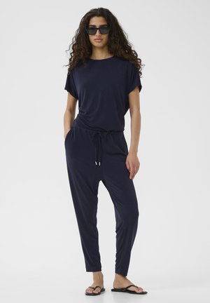 Combinaison bleu marine à manches courtes, taille élastique et cordon de serrage réglable. Tissu lisse, jambes ajustées, portée avec des tongs noires.
