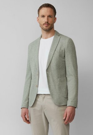 Hellgrau strukturiertes Blazer mit Reverskragen, zwei Knöpfen und einer Tasche. Über einem weißen Hemd getragen und kombiniert mit beigen Hosen.