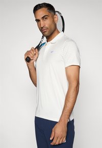 Balta polo krekls ar teksturētu punktiņu rakstu, klasiskā apkakle un sānu slīpsavienojumi, kopā ar tumši ziliem šortiem. Tur rokās tenisa raketi.