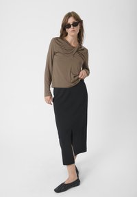 Haut marron à manches longues avec un design drapé, associé à une jupe noire mi-longue avec une fente sur le côté ; le mannequin porte des chaussures plates noires.