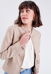 Beżowa skórzana kurtka z zapięciem na zamek błyskawiczny, klapką zapinaną na napy oraz prążkowanymi akcentami. Model trzyma kurtkę, mając na sobie białą koszulę i jasnoniebieskie dżinsy.