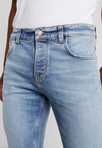 Ljust blå jeans i denim med en figurnära design, med dragkedja, kopparnitar och en liten logotyp på midjebandet.