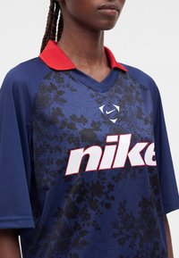 Άτομο που φορά μπλε πουκάμισο Nike με κόκκινο γιακά και μαύρο λουλουδάτο σχέδιο, δείχνει το πάνω μέρος του κορμού και πλεξουδάκια σε απλό γκρι φόντο.