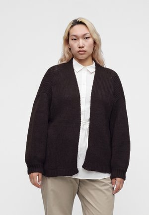 Person med langt blondt hår iført en mørk strikket cardigan over en hvid skjorte med knapper og beige bukser, stående mod en ensfarvet baggrund.