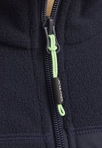 Chaqueta de forro polar azul marino con una cremallera prominente, con un tirador verde y una etiqueta negra que dice "JACK&JONES". Textura suave y detalles cosidos.