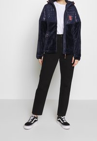 Henkilö, jolla on yllään tummansininen fleecetakki ja tikattu takki, valkoinen t-paita, mustat housut ja mustavalkoiset lenkkarit, seisoo harmaalla lattialla.