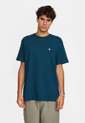Jonge man met kort krullend haar die een donker teal kleurig t-shirt draagt met een klein wit vogel-logo en lichtbeige broek.
