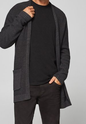 Personne portant un cardigan gris foncé ouvert à l’avant avec une poche, une chemise noire et un pantalon sombre, ajustant le col du cardigan d’une main.