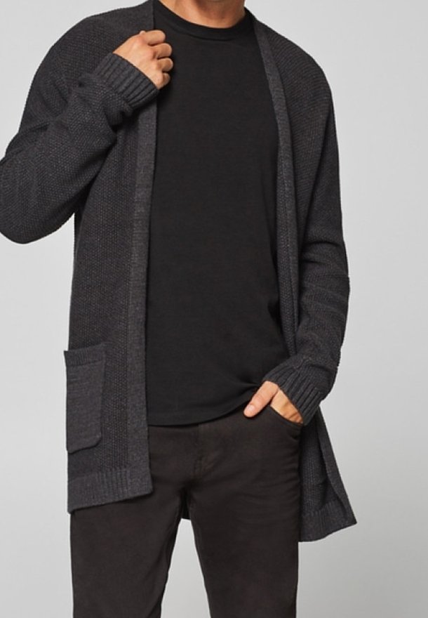 Personne portant un cardigan gris foncé ouvert à l’avant avec une poche, une chemise noire et un pantalon sombre, ajustant le col du cardigan d’une main.
