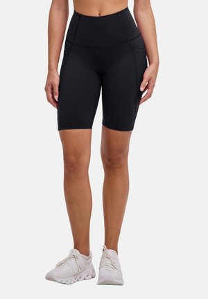 Frau trägt schwarze Biker-Shorts mit hoher Taille und Seitentaschen sowie weiße Sportschuhe, steht vor einem einfarbigen Hintergrund.