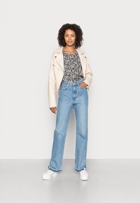Jaqueta de couro creme, blusa com padrão floral, jeans de perna larga azul claro e ténis brancos. Cabelo encaracolado emoldura o rosto.