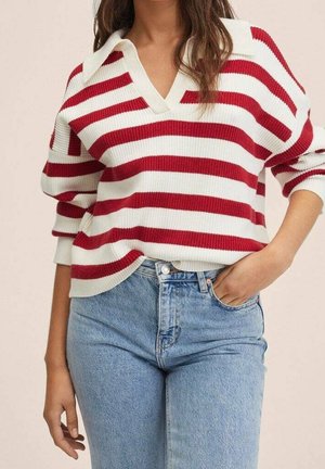 Femme portant un pull en maille blanc et rouge à rayures avec un col en V et un jean taille haute bleu clair, une main dans la poche.