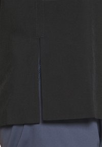 Vestuário preto com uma bainha fendida, apresentando uma textura suave. Por baixo, uma camada azul-marinho é parcialmente visível, acrescentando profundidade ao design.