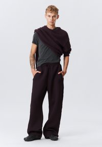 Dunkelgraues T-Shirt, das unter einem kastanienbraunen gestrickten Schal getragen wird, kombiniert mit lockeren schwarzen Sweatpants und strukturierten schwarzen Schuhen.